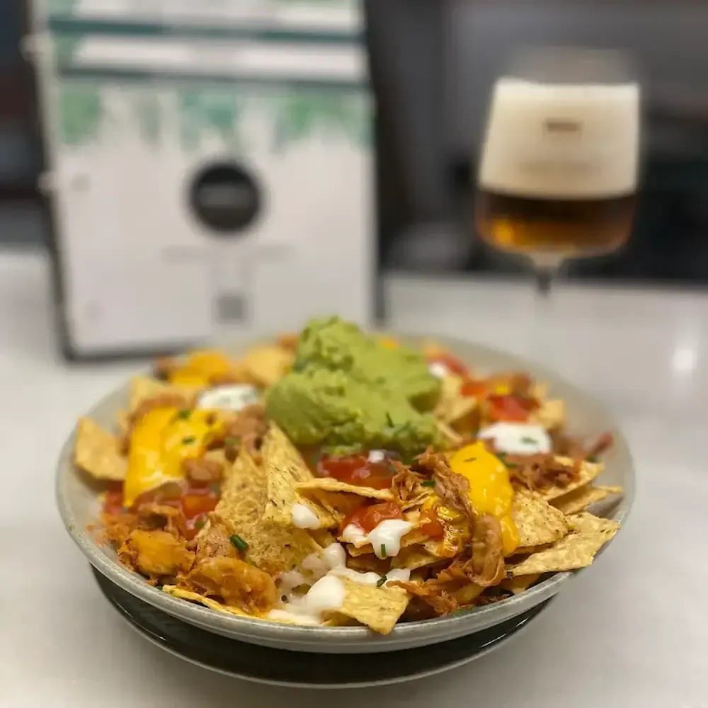 Nachos cubiertos con queso, salsa, guacamole y crema agria, con una cerveza de fondo.