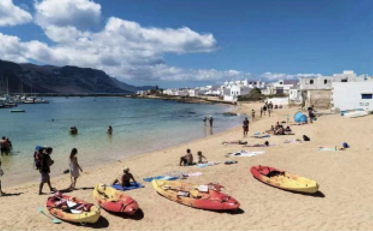 Playa soleada con kayaks en la arena, bañistas en aguas azules tranquilas y edificios blancos a lo largo de la costa.