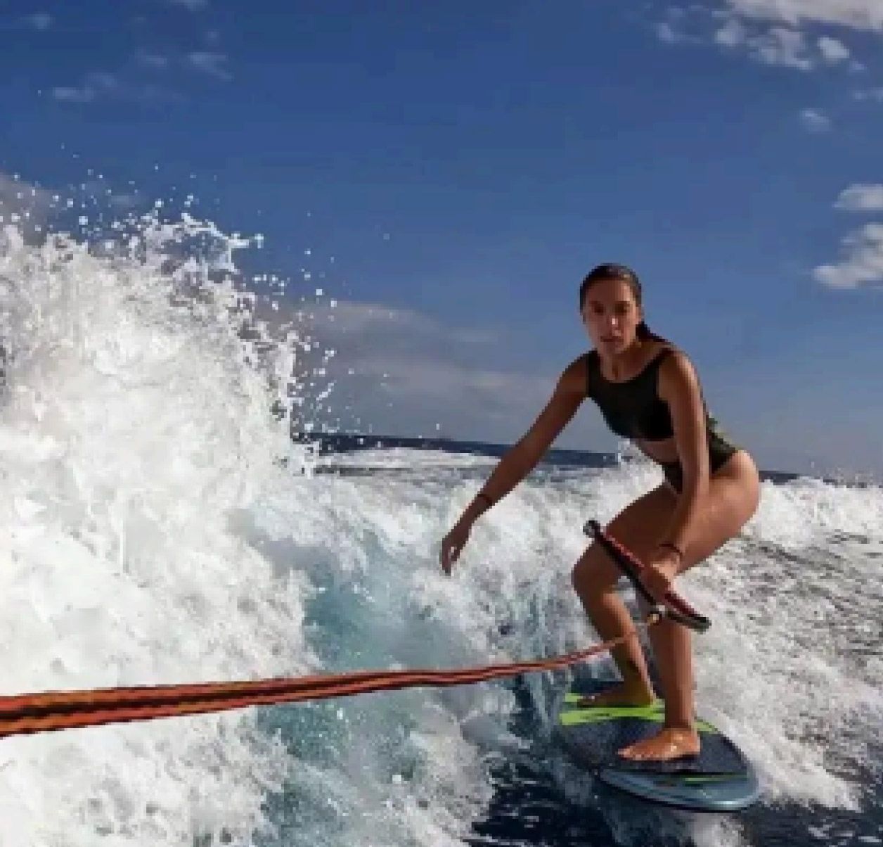 Una persona practica wakesurf detrás de una lancha en aguas azules, sujetando una cuerda de remolque mientras las olas salpican a su alrededor.