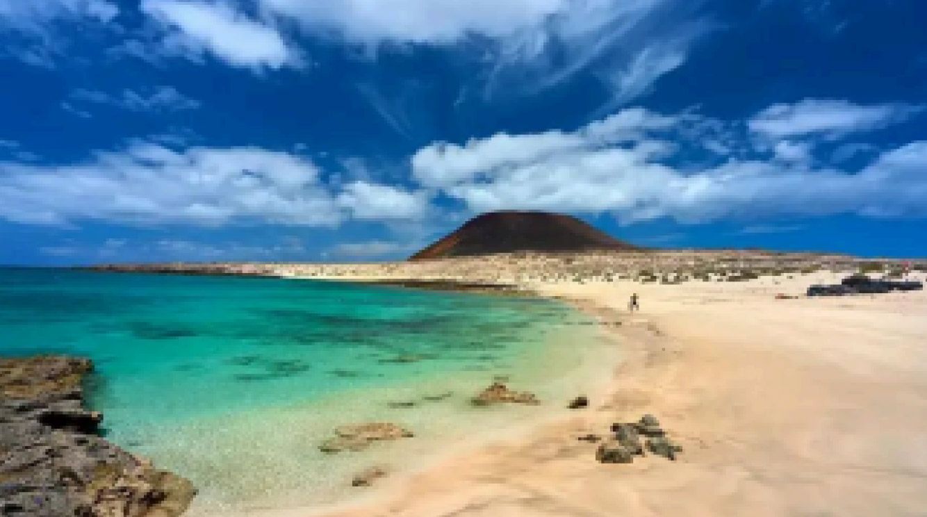 Laguna turquesa y playa de arena con costa rocosa, cielo azul y una pequeña colina volcánica en la distancia.