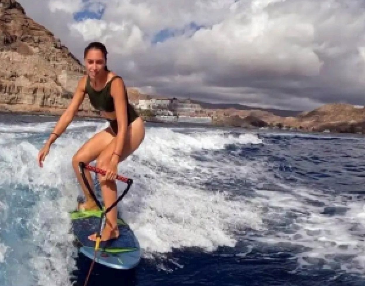 Mujer surfeando sobre una ola cerca de una costa rocosa bajo un cielo nublado.