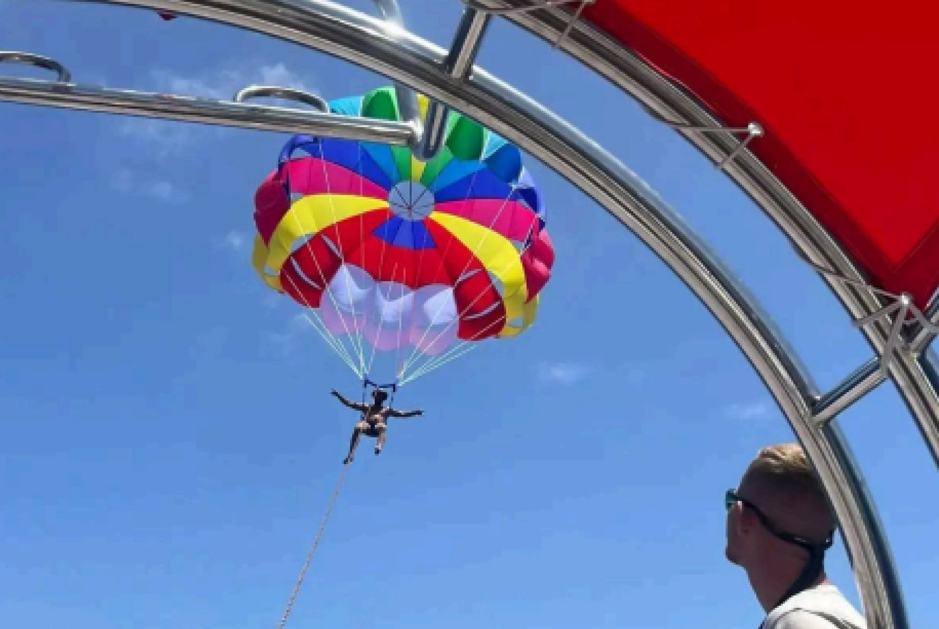 Un colorido parapente con una persona colgando debajo, con un cielo azul de fondo y enmarcado por el aparejo de un barco.