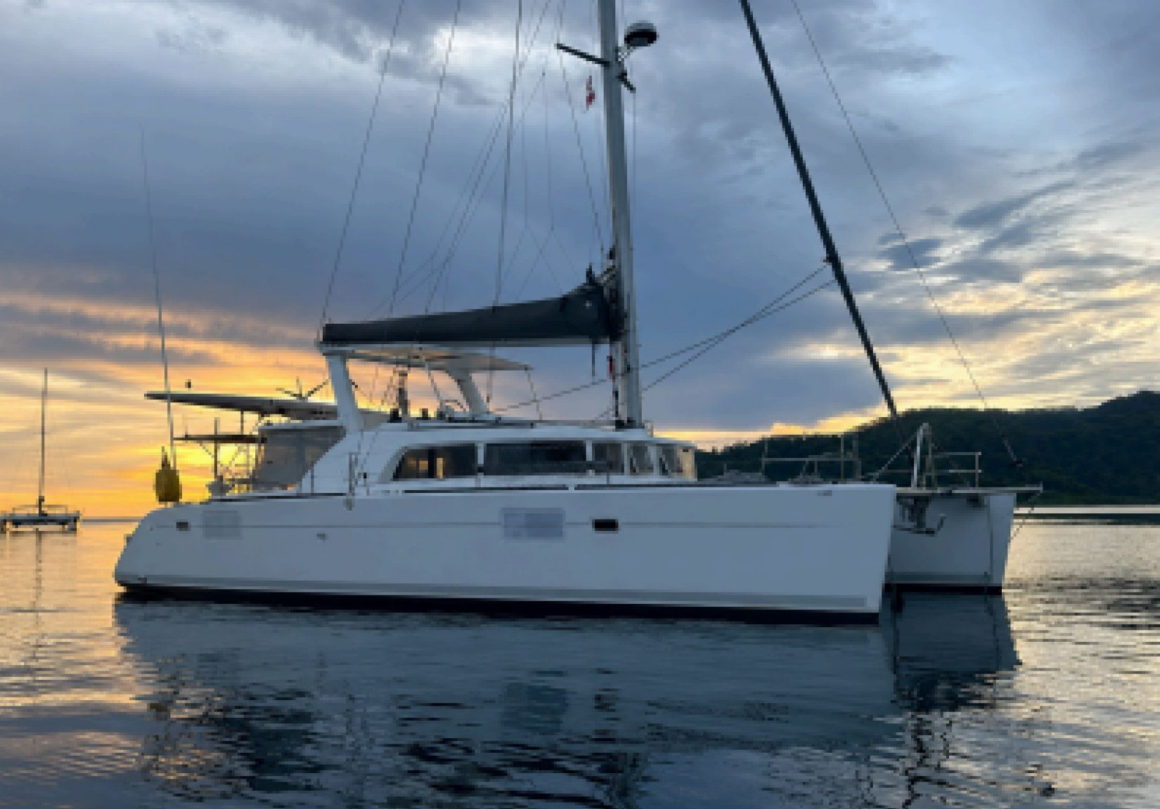 Velero catamarán blanco al atardecer sobre aguas tranquilas con cielo nublado.