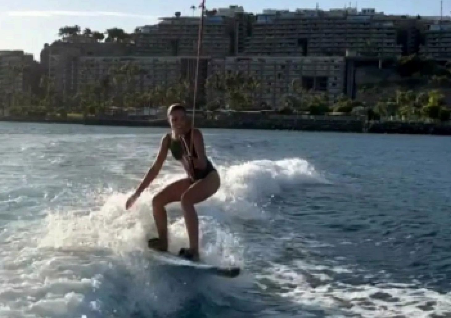Una persona practica wakesurf en una tabla detrás de una lancha en aguas abiertas, con edificios costeros al fondo.