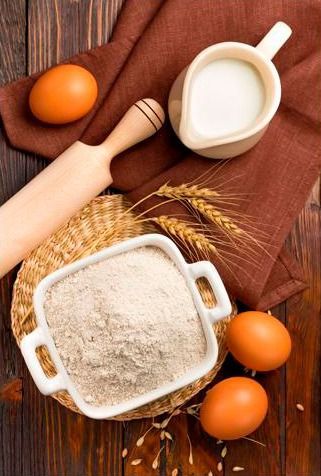 Un bol con harina, huevos, leche y un rodillo sobre una mesa de madera.