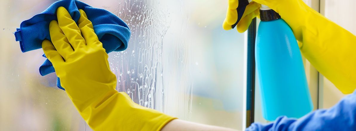 Person sprüht Glasreiniger auf Fenster, die andere Hand hält ein Microfasertuch