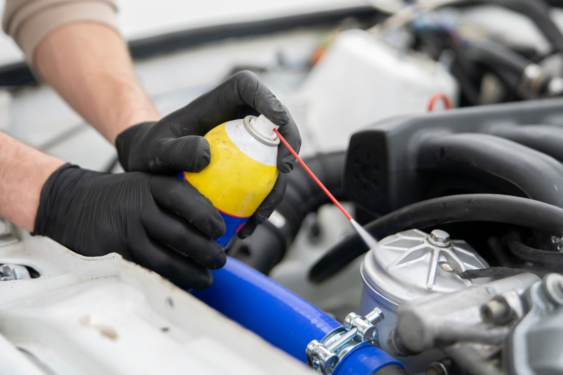 Une personne vaporise un moteur de voiture avec une bombe aérosol jaune et bleu, portant des gants noirs.