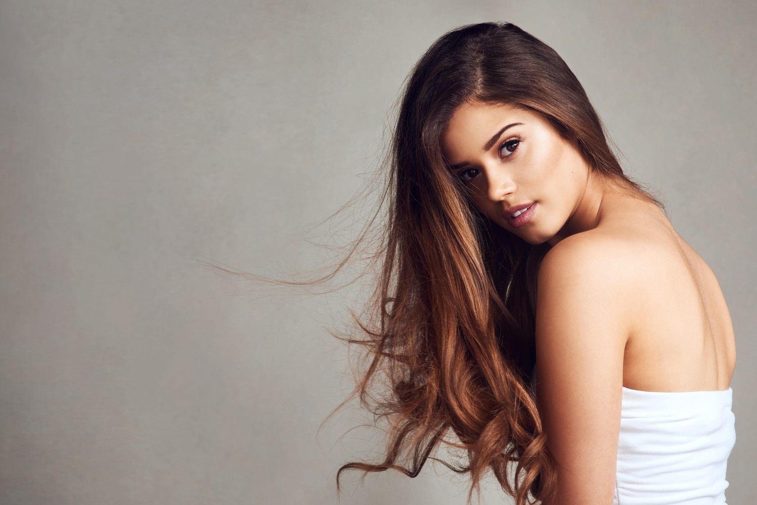 Mujer con cabello castaño largo con un top blanco sin tirantes sobre un fondo gris.