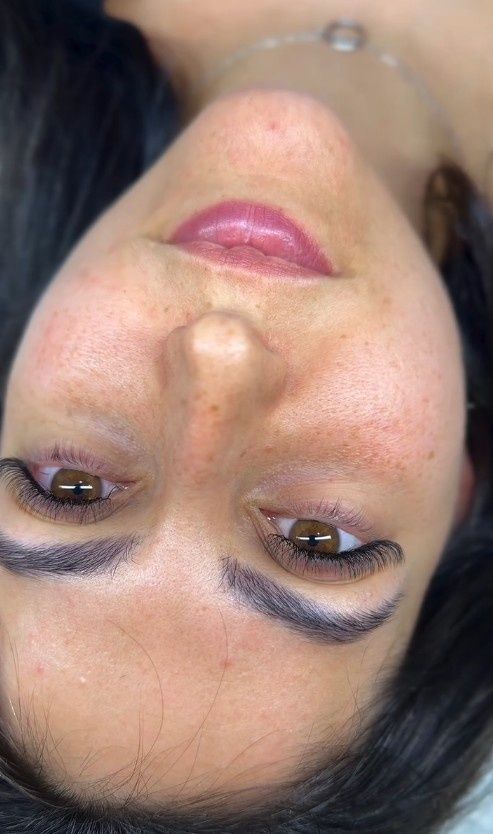 Primer plano de un rostro invertido con cabello oscuro, cejas arqueadas y lápiz labial rosa sobre un fondo claro.