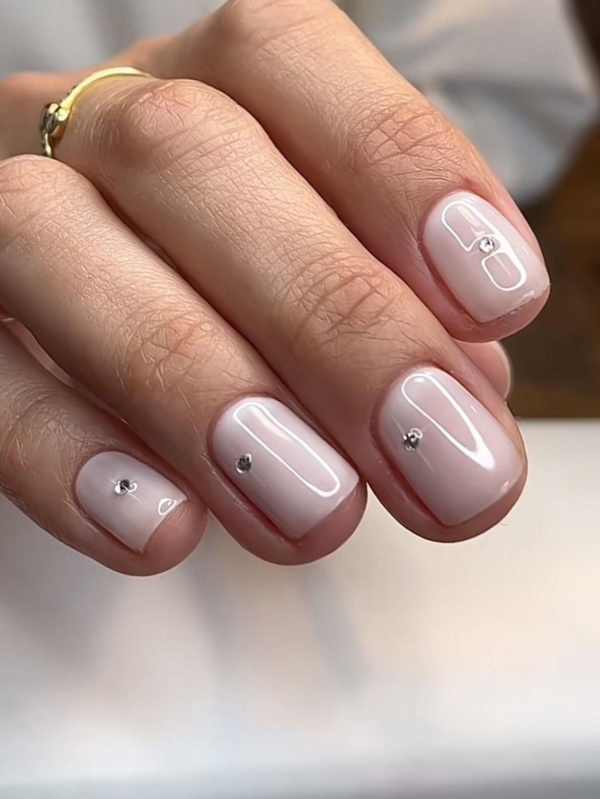Mano con uñas cortas de color rosa pálido y pequeños remaches plateados, mostrada en primer plano.
