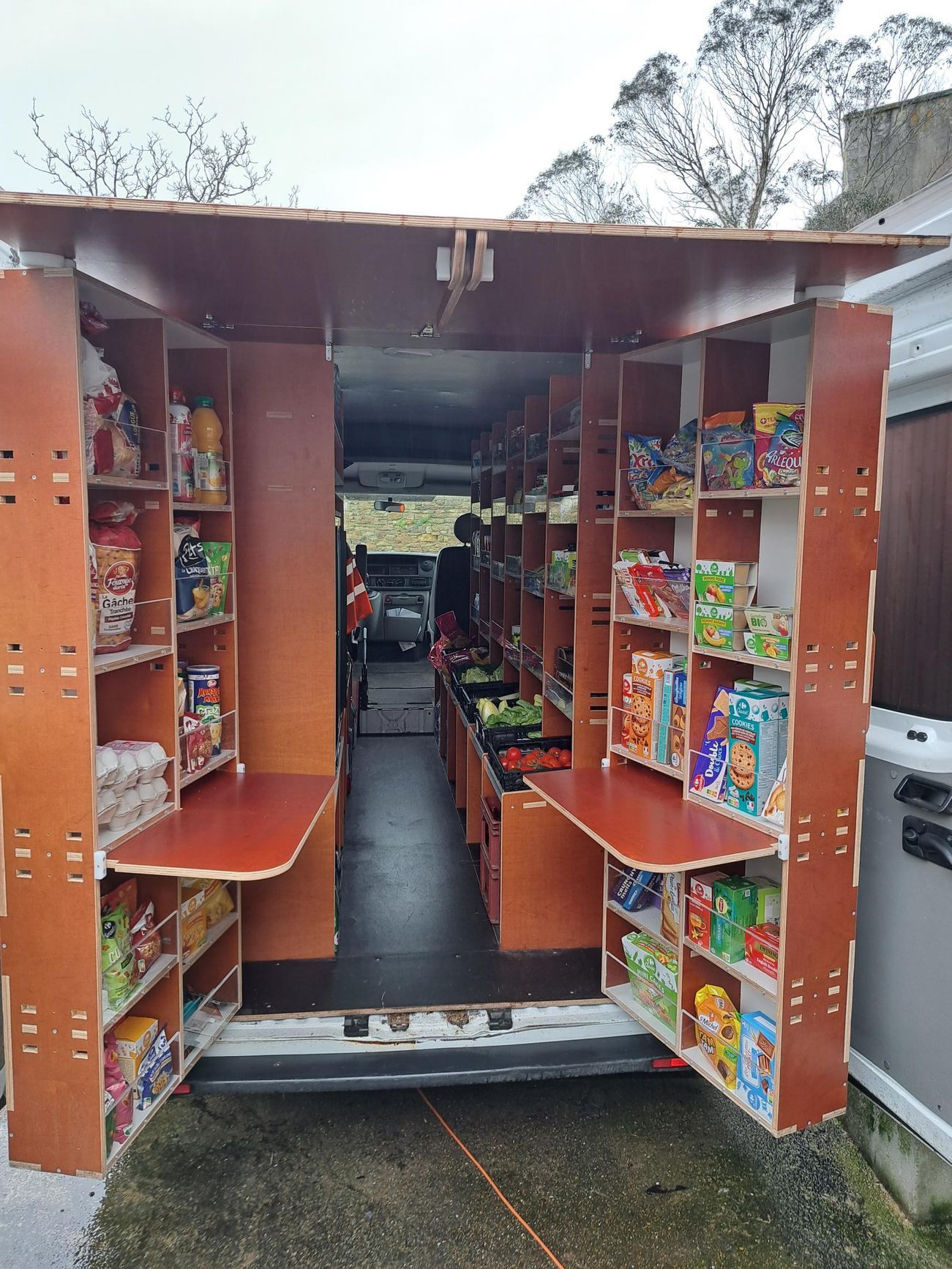 Camionnette-épicerie mobile avec étagères ouvertes remplies de nourriture. Deux comptoirs rabattables.