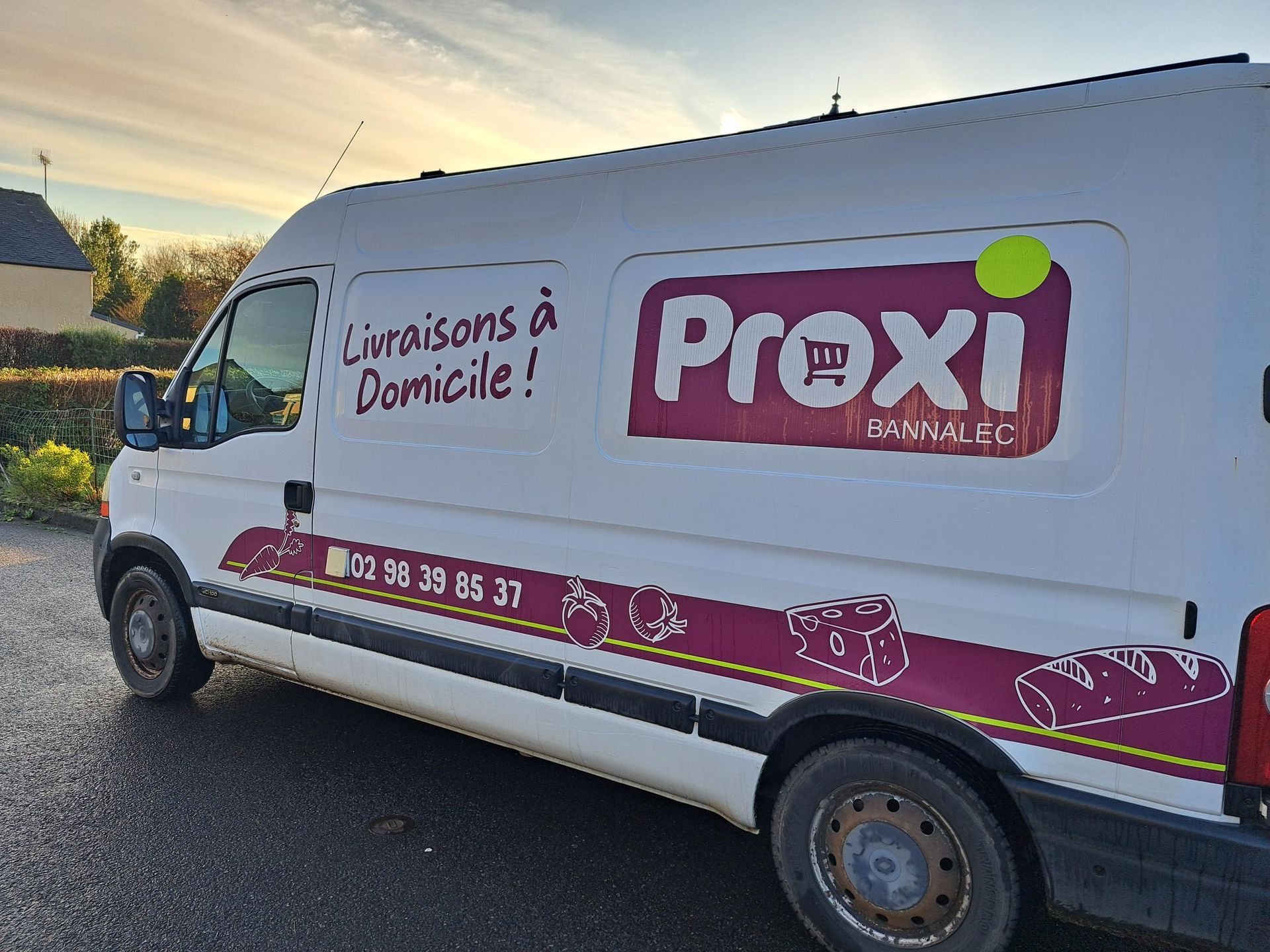 Camionnette de livraison blanche avec le logo Proxi et l'inscription « Livraisons à Domicile ! », garée à l'extérieur.