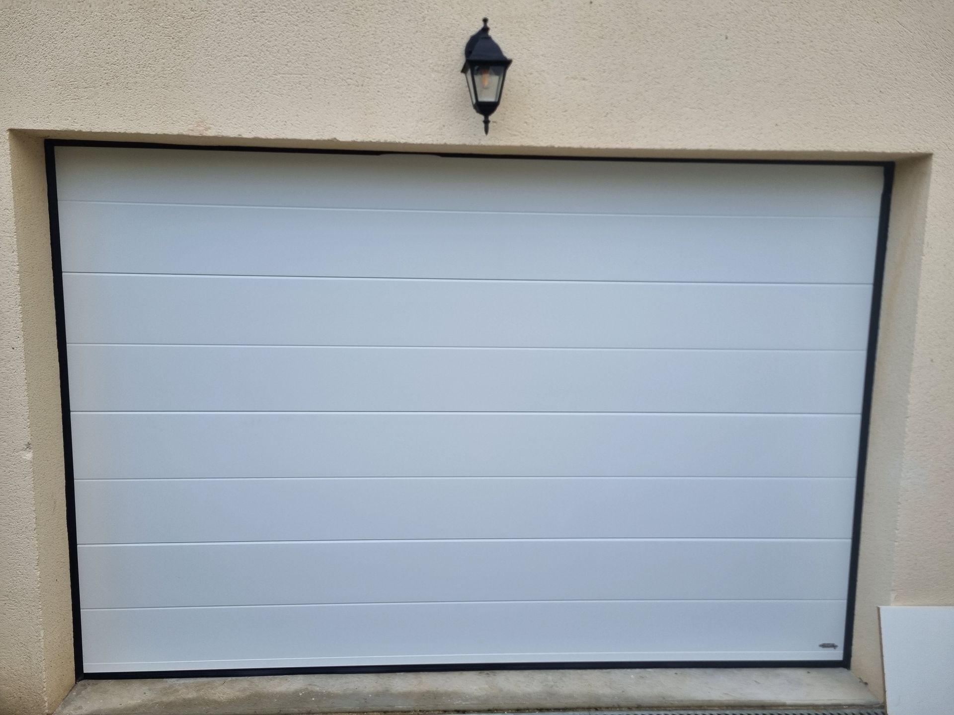 Porte de garage blanche à rayures horizontales, encadrée de noir, sous un mur beige, et une lumière noire.