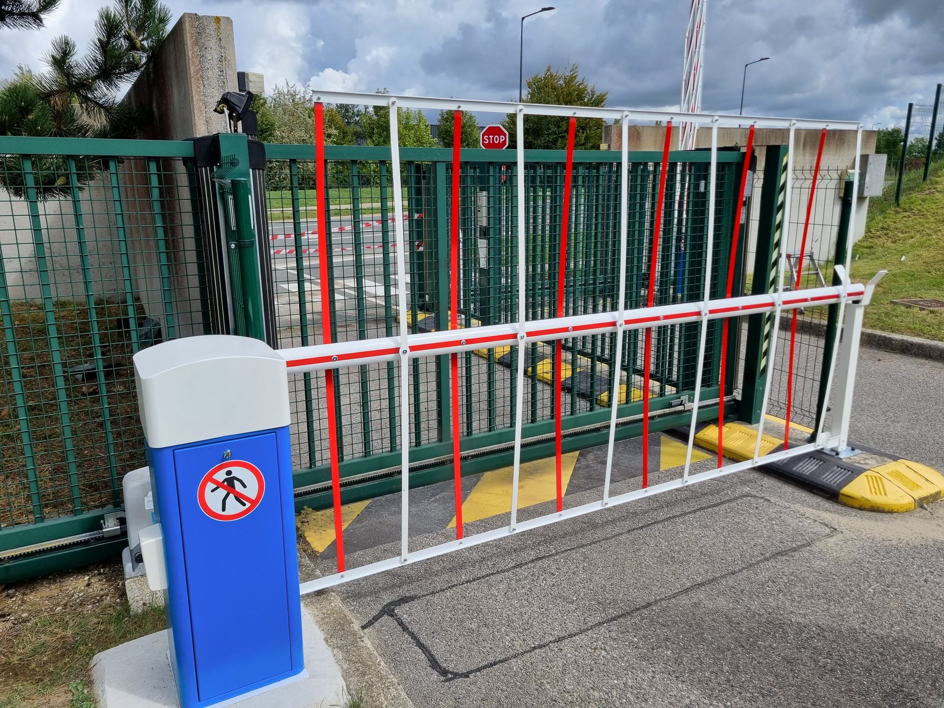 Une barrière à l'entrée. La barrière est blanche à rayures rouges. Un panneau de commande bleu se trouve à gauche.