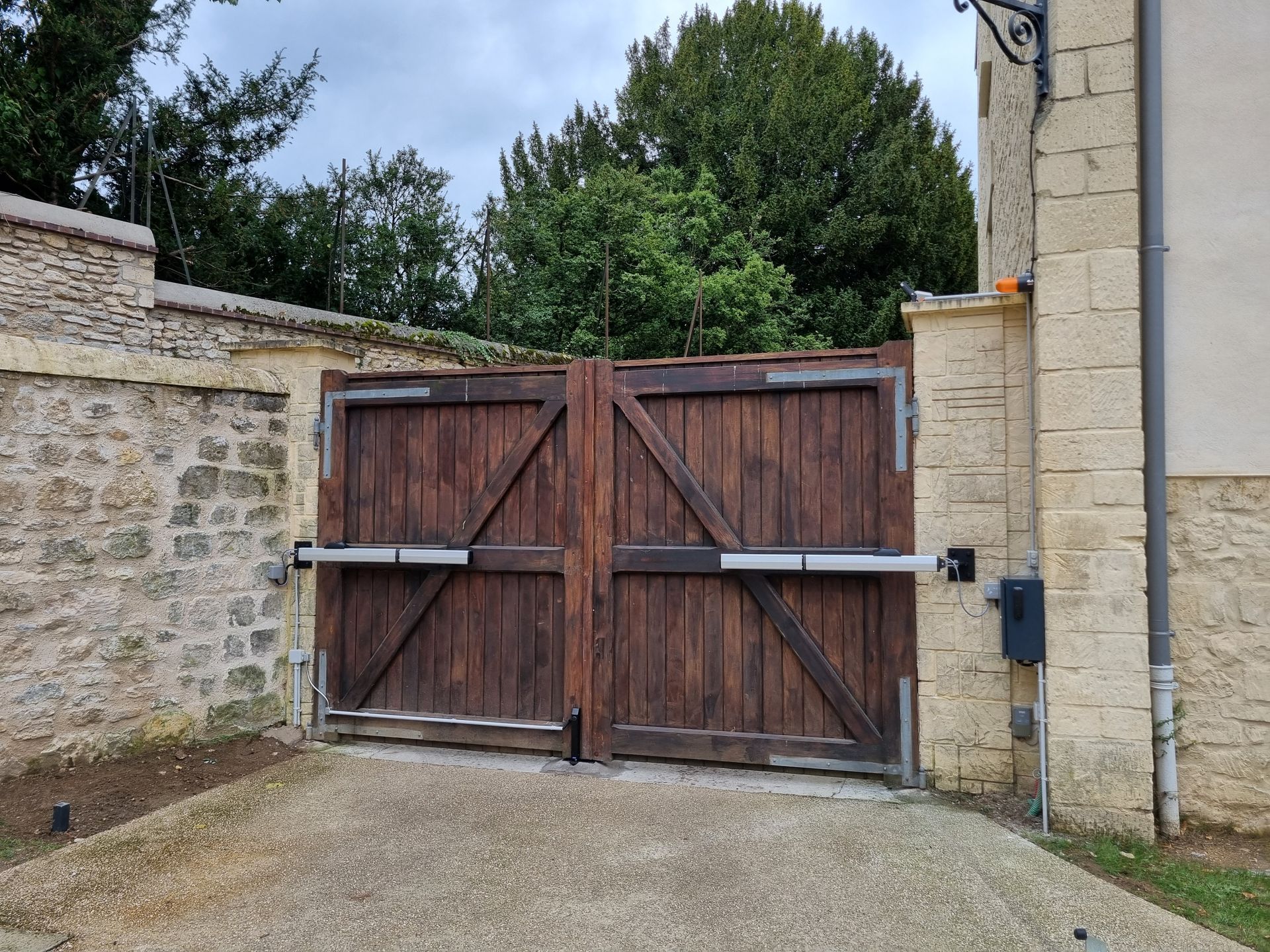 Portail double en bois avec bras motorisés, intégré à un mur en pierre. Allée gravillonnée.