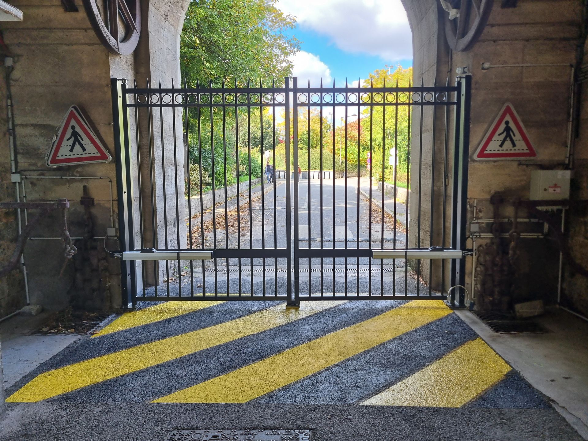 Portail métallique noir avec panneaux de passage piéton et bandes de signalisation jaunes et noires.
