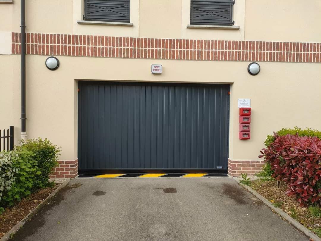 Entrée de garage avec porte grise fermée, éclairage, garniture en briques et panneau d'avertissement à rayures noires et jaunes.
