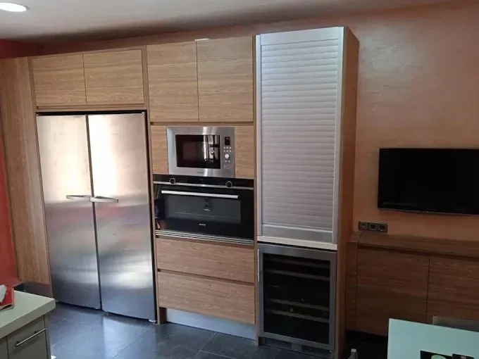 Un mueble de cocina con acabado en madera que incluye frigorífico de acero inoxidable, microondas, horno empotrado.