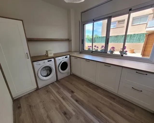 Un cuarto de lavado con armarios blancos, suelo que imita la madera, dos lavadoras y una gran ventana con vistas al exterior.