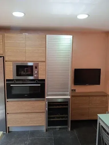 Una pared de la cocina con armarios de madera, microondas integrado, horno, vinoteca y televisor montado en la pared.