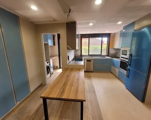 Una cocina moderna con armarios azules, suelo de madera clara, una isla de madera y una zona de servicio independiente.