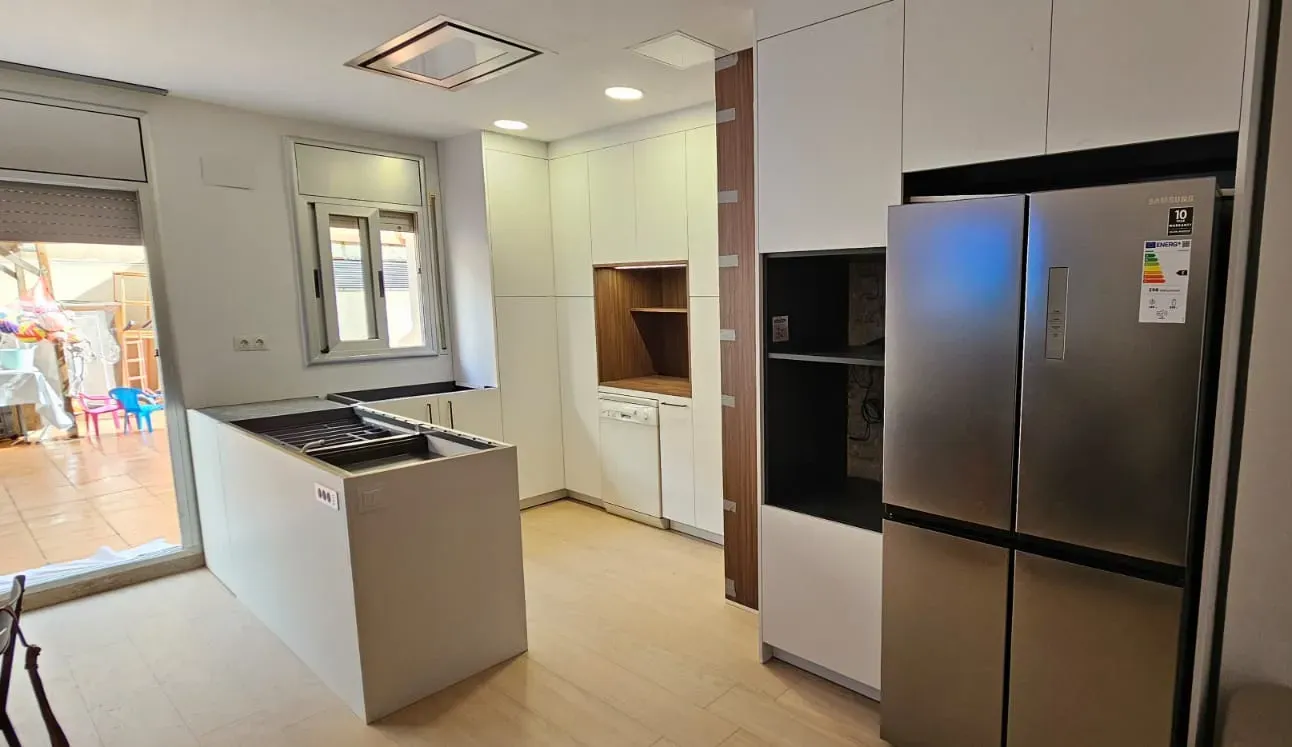 Cocina moderna con refrigerador de acero inoxidable, gabinetes blancos, una isla central con estufa y pisos de madera clara.