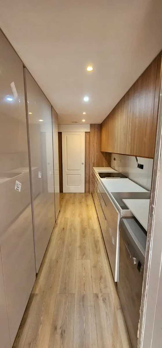 Una cocina estrecha y moderna en un pasillo, con armarios en tono madera, paneles brillantes a la izquierda.