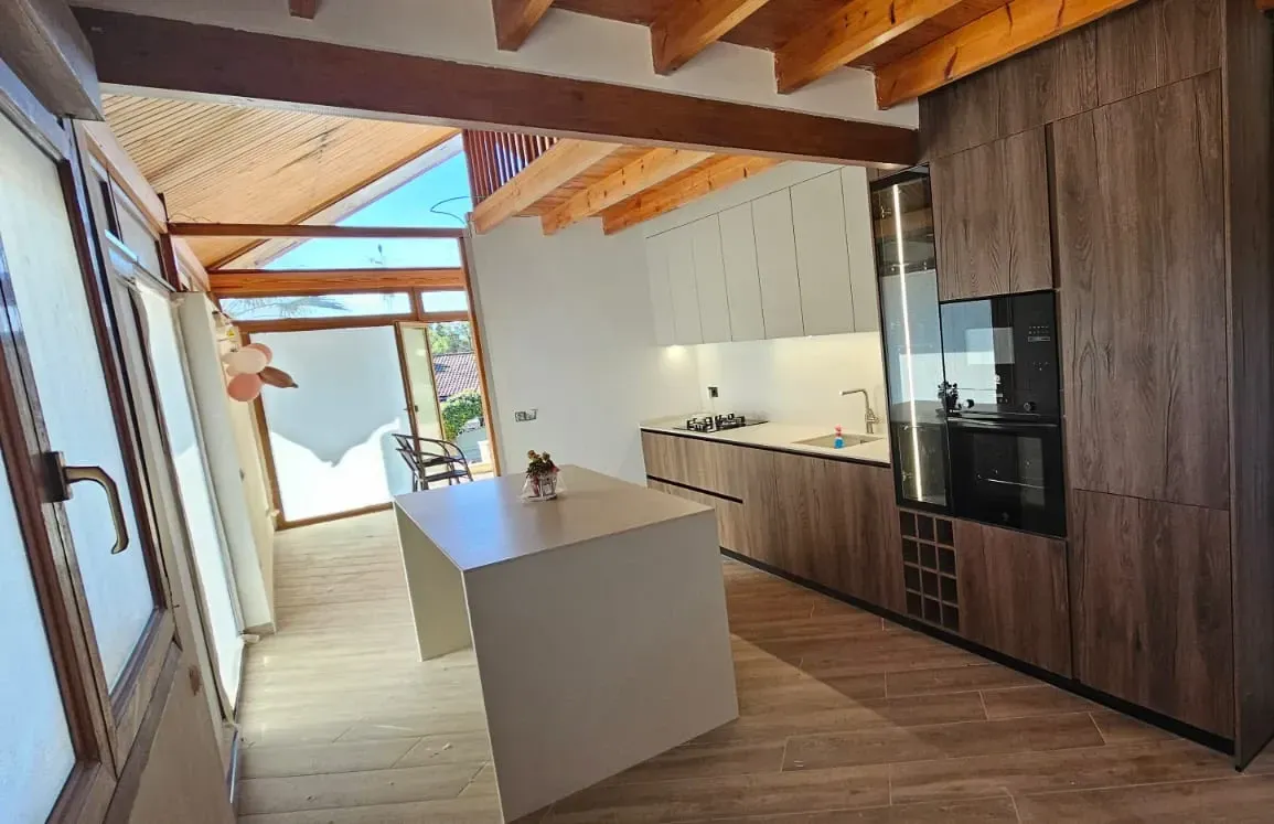 Cocina con armarios de madera oscura, armarios superiores blancos, una isla central y una zona acristalada con vigas.