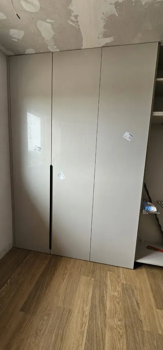 Un armario de tres puertas de color claro instalado en una habitación con suelo de madera y techo sin terminar.