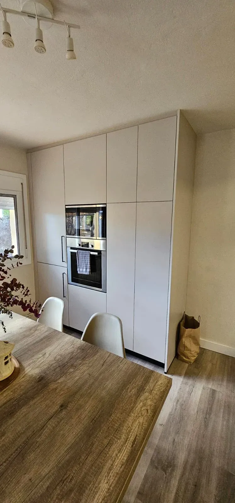 Un mueble de cocina blanco que va desde el suelo hasta el techo, con horno, junto a una mesa y un suelo de madera.