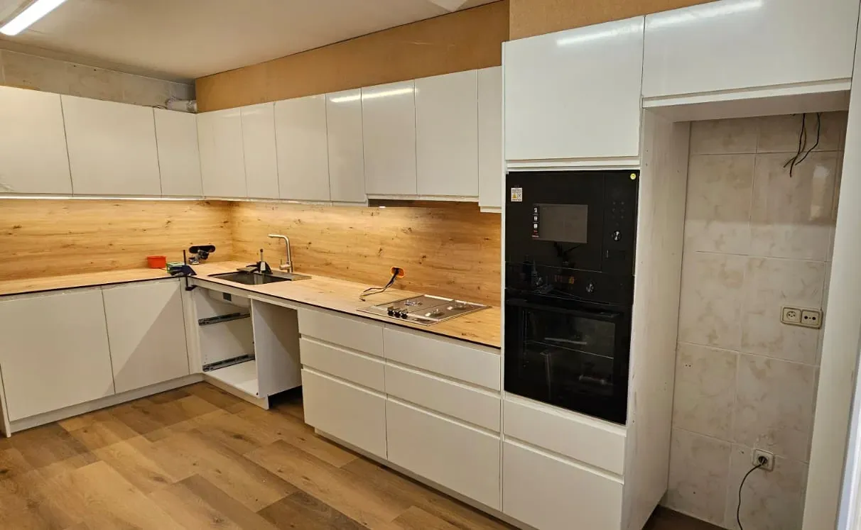 Una cocina con armarios blancos, encimeras de madera, un horno empotrado negro y espacio preparado para un frigorífico.