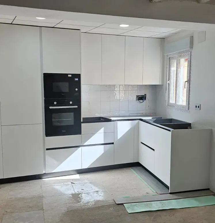 Una cocina con armarios blancos, horno y microondas empotrados de color negro y un fregadero negro debajo de una ventana.