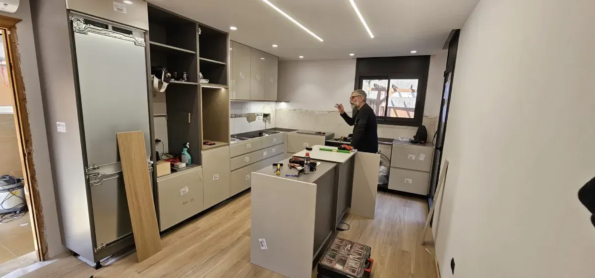 Una persona instalando armarios de cocina en una cocina moderna de madera clara, con armarios grises y una isla.