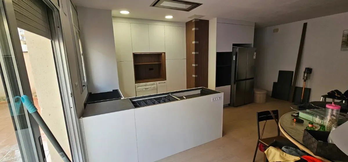 Una cocina sin terminar con una isla blanca, armarios blancos, un nicho en tono madera y un frigorífico de acero inoxidable.