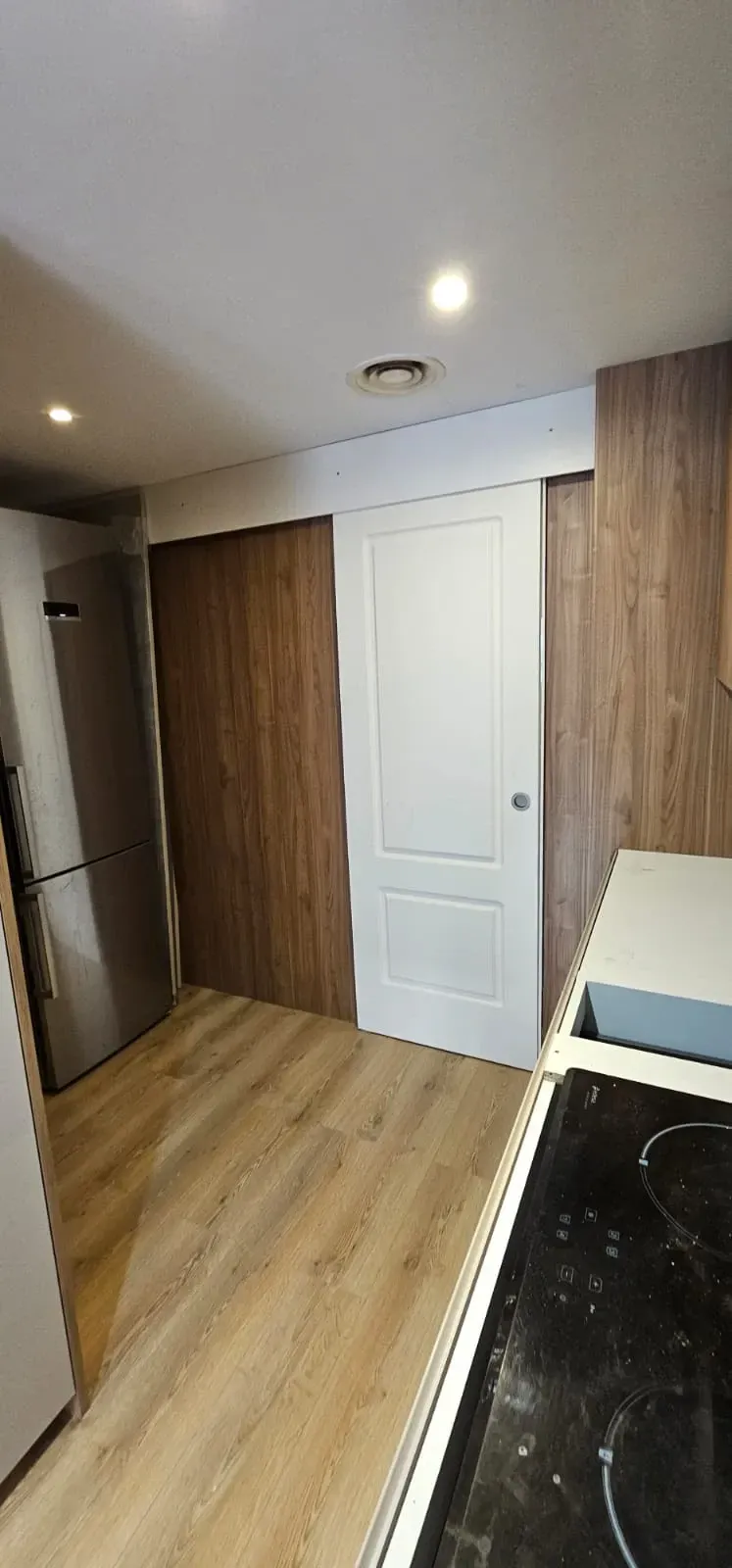 Interior de cocina con paredes revestidas de madera, una puerta blanca, un frigorífico de acero inoxidable.