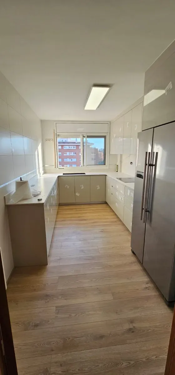 Una cocina alargada y estrecha, con suelo de madera, armarios y encimeras blancas y un frigorífco de acero inoxidable.