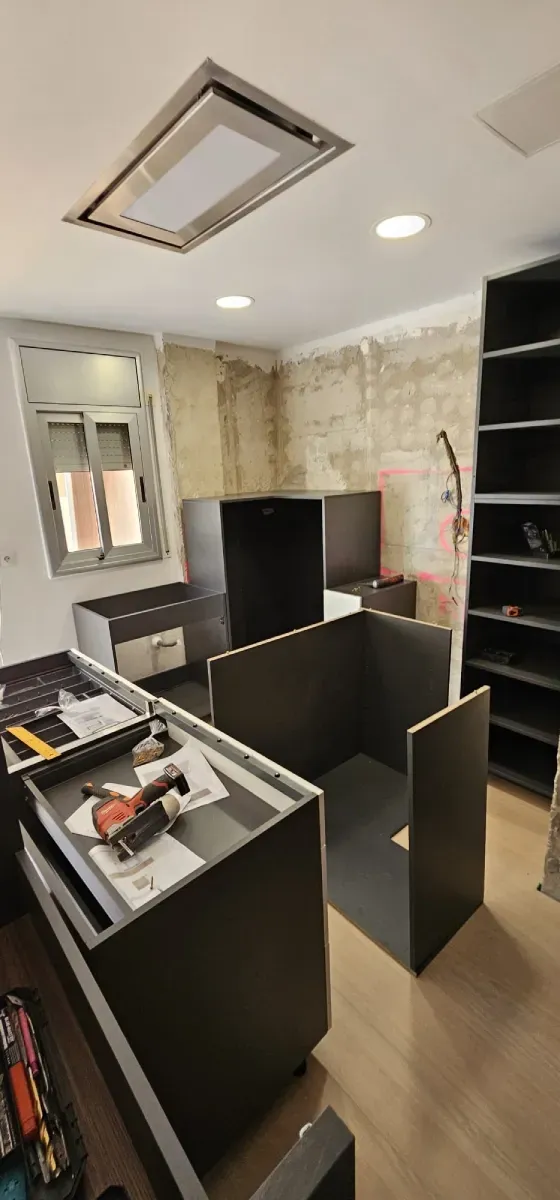 Una cocina en construcción con armarios oscuros, una estantería abierta y una pared sin terminar.