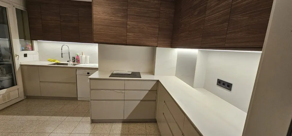 Una cocina con armarios inferiores blancos, armarios superiores, encimera blanca e iluminación bajo los armarios.