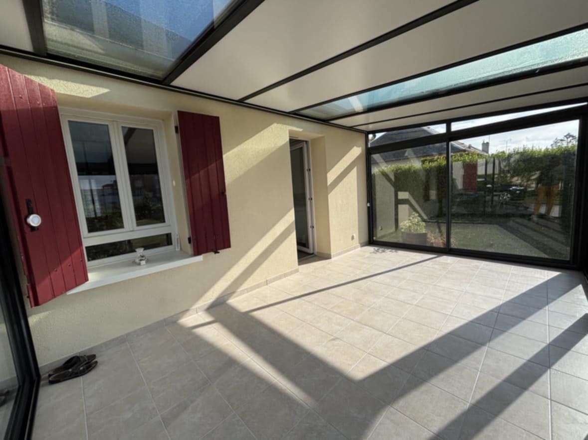 Pièce intérieure ensoleillée avec verrière et portes coulissantes. Fenêtres avec volets rouges. Sol carrelé beige.