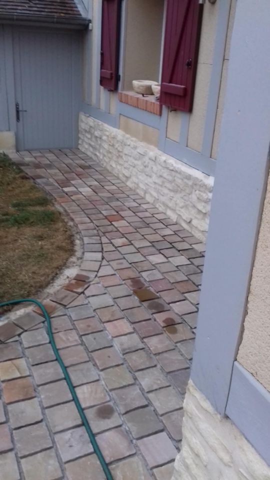 Un chemin en briques serpente le long d'une maison, avec un revêtement en pierre et des volets rouges.