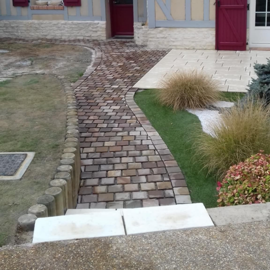 Chemin de briques menant à une maison avec porte rouge, bordée d'herbe et de petits buissons.