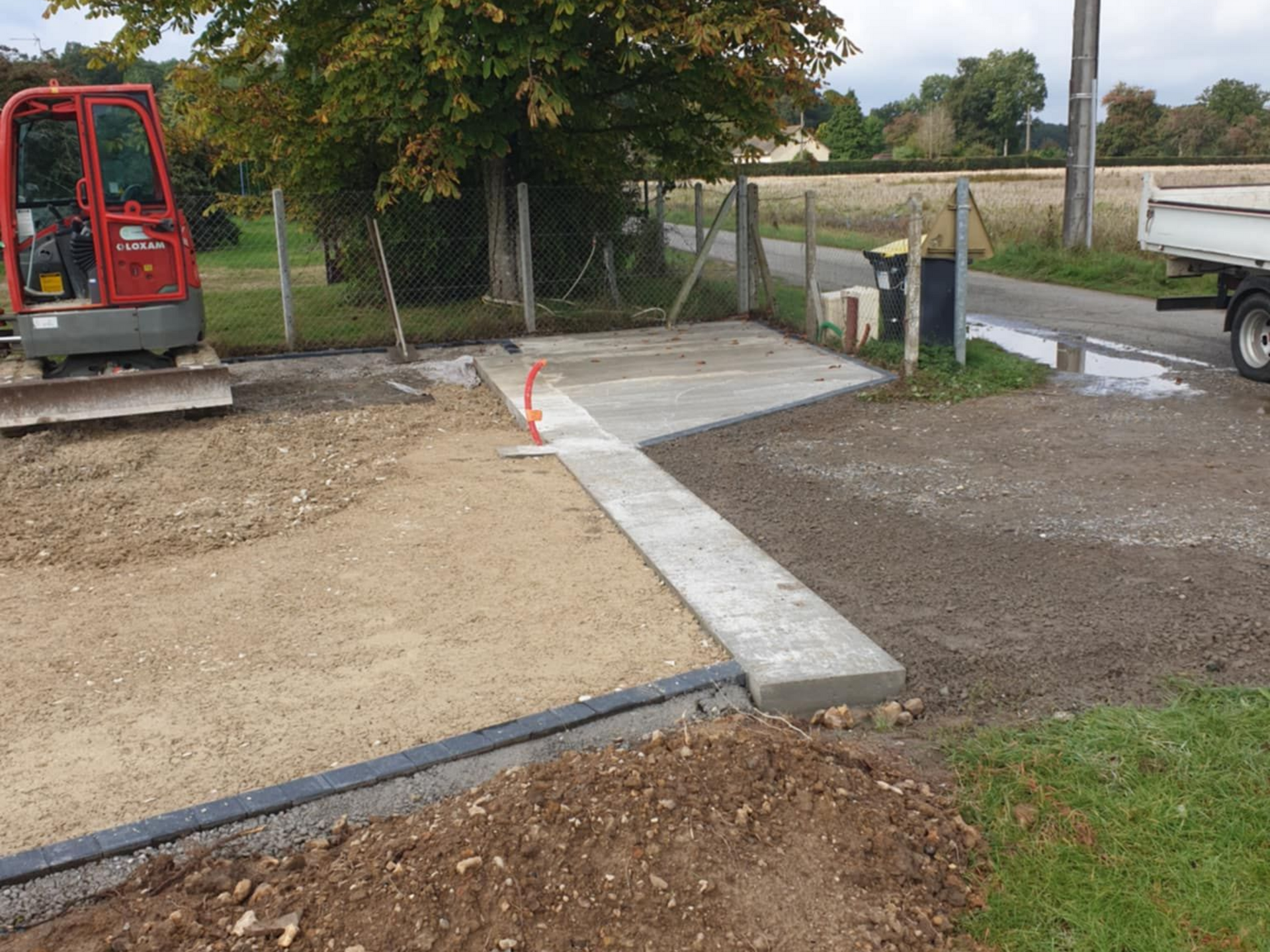 Chantier de construction avec petite excavatrice, allée en béton, gravier et clôture.