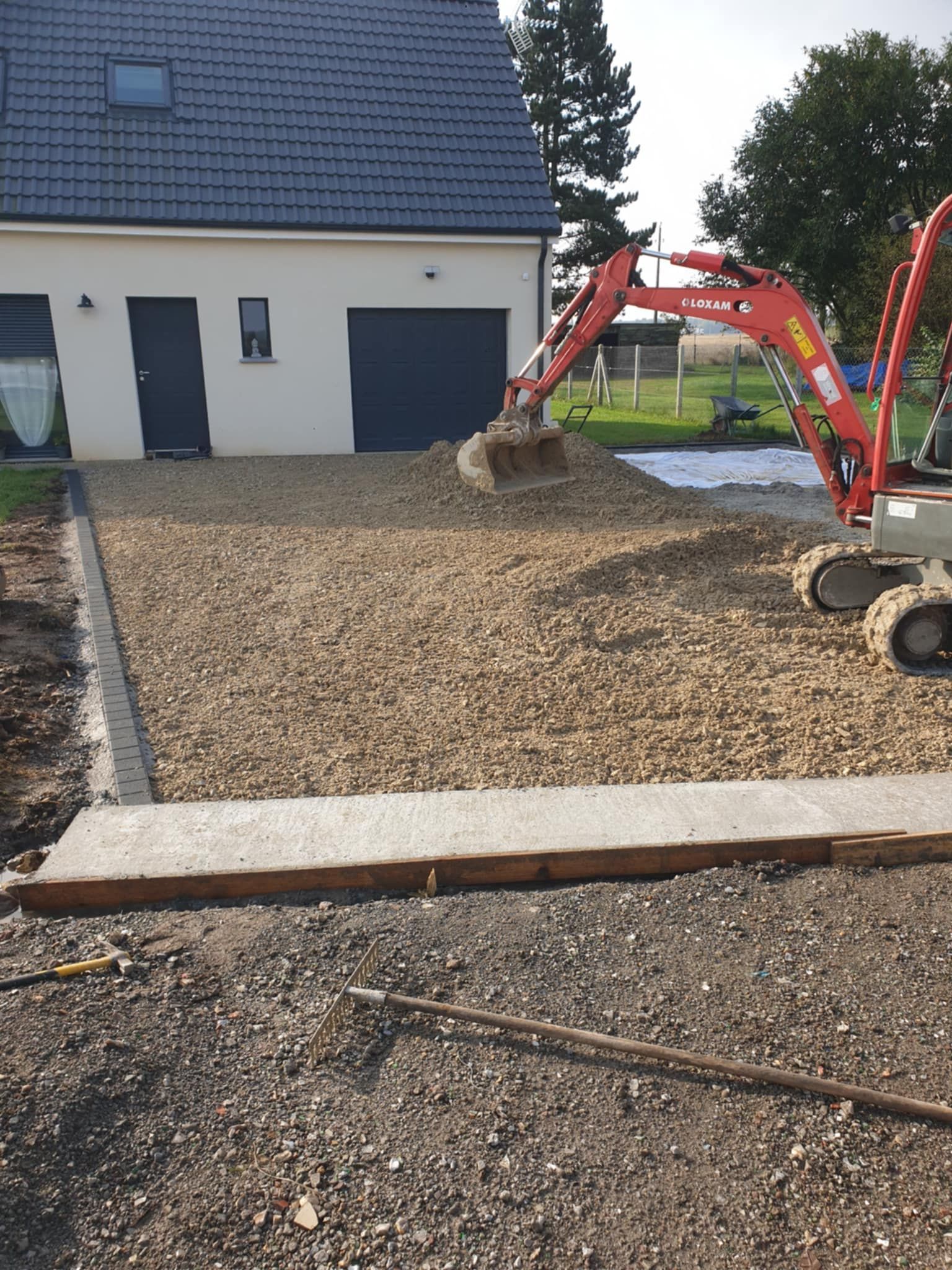 Une petite excavatrice répand du gravier sur une allée en construction près d'une maison blanche.