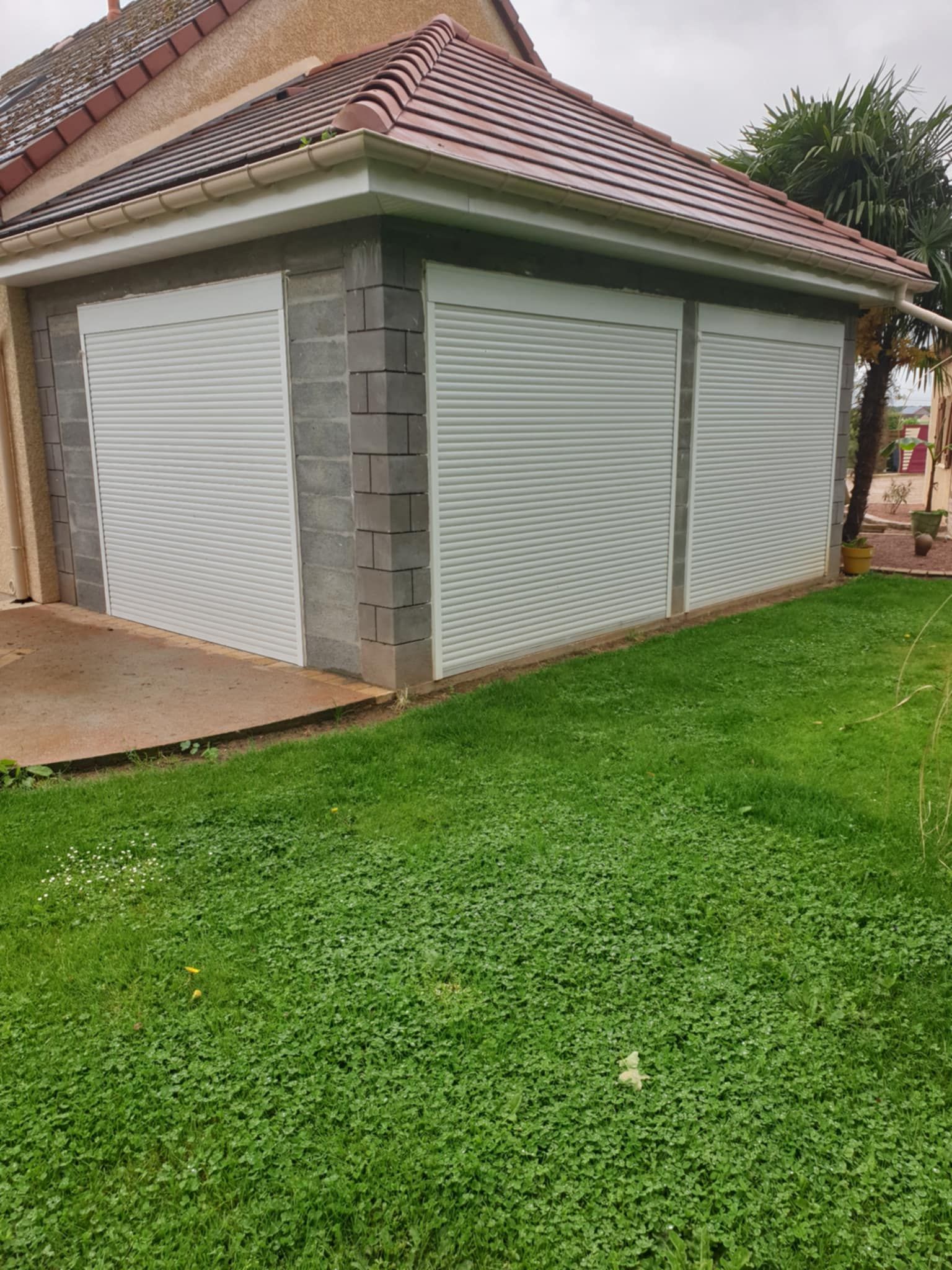Portes de garage en tôle ondulée blanche sur une structure en parpaings avec un toit en tuiles rouges, posées sur de l'herbe verte.