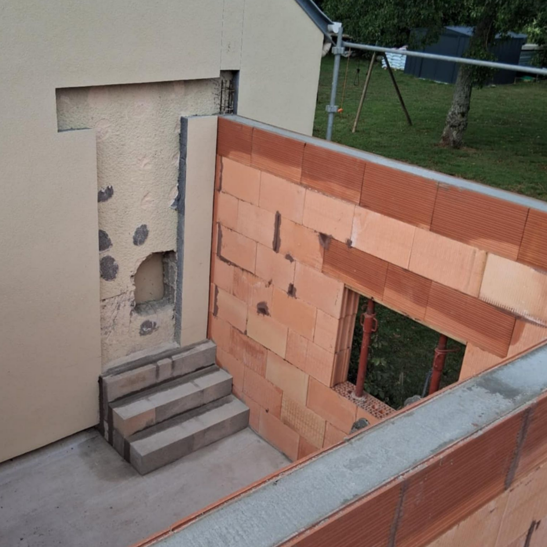 Chantier avec mur de briques rouges partiellement construit, petites marches menant à une entrée en retrait.