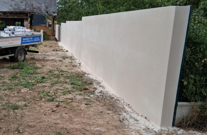 Mur beige fraîchement enduit à l'extérieur, le long d'une bordure de gravier et d'herbe, avec un camion et des arbres en arrière-plan.