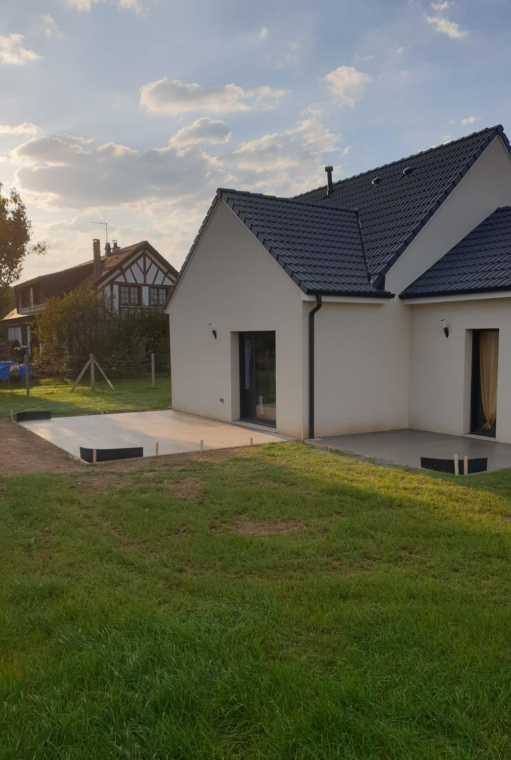 Une maison de couleur claire avec un toit sombre et un patio sur une pelouse herbeuse sous un ciel partiellement nuageux.