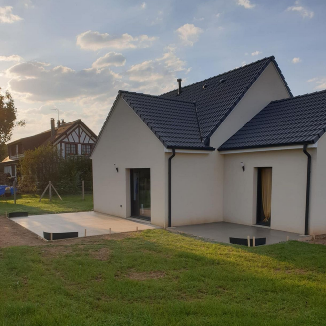 Maison blanche avec toit sombre, terrasse en béton et pelouse verte par une journée ensoleillée.