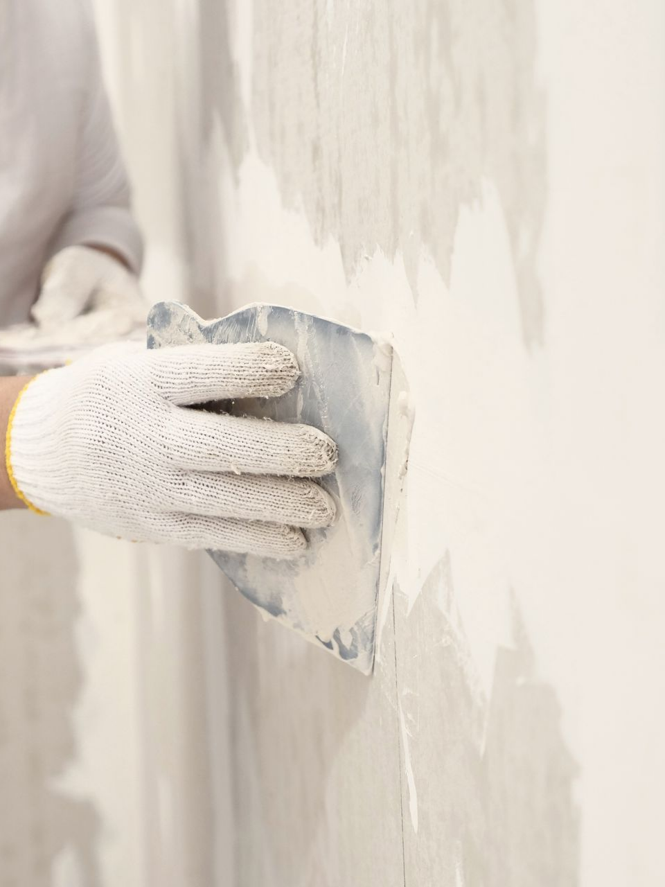 Une personne portant des gants utilise un couteau à mastic pour appliquer du mastic sur un mur blanc.