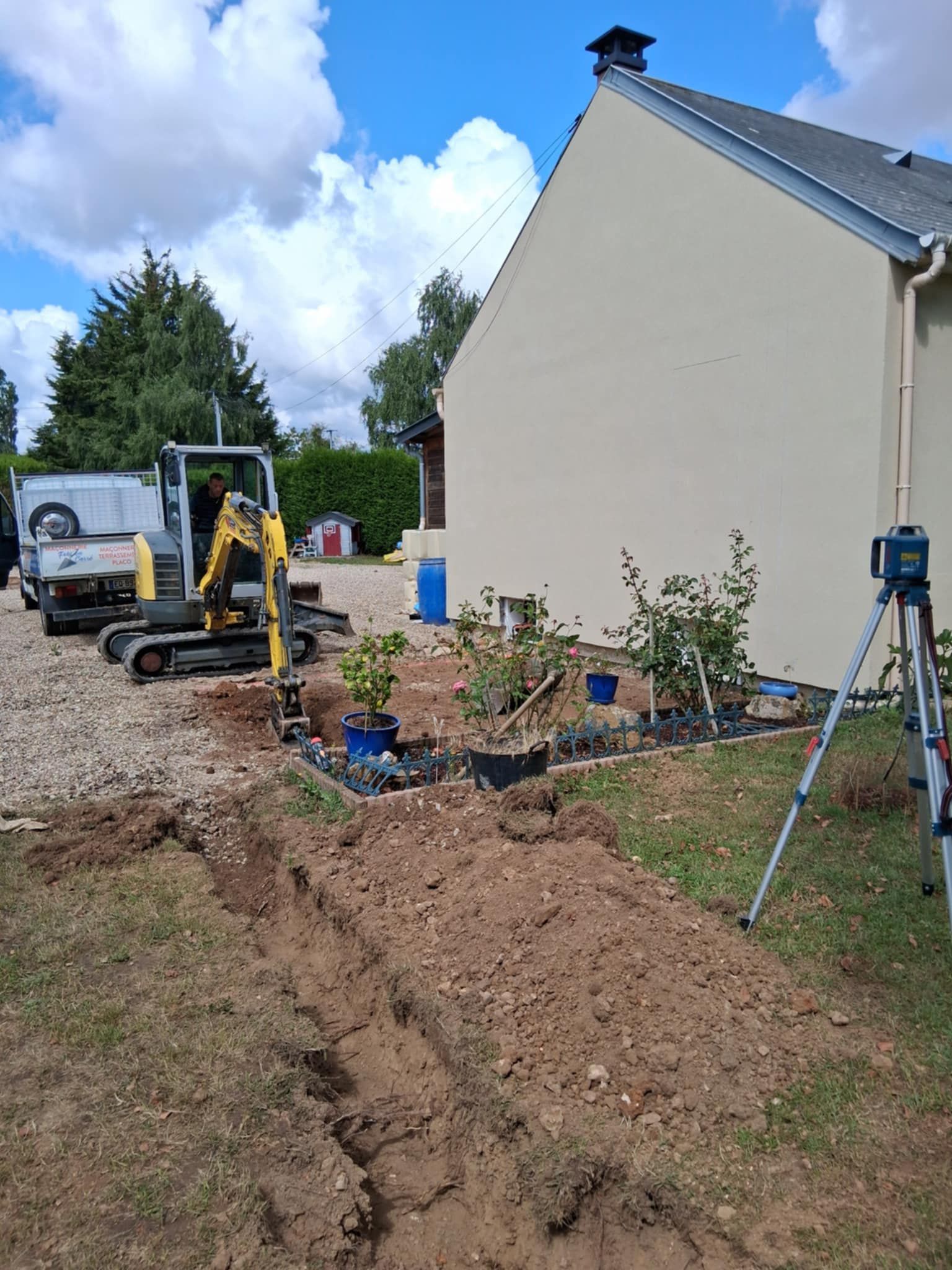 Chantier de construction avec une excavatrice creusant une tranchée à côté d'une maison beige par une journée ensoleillée.
